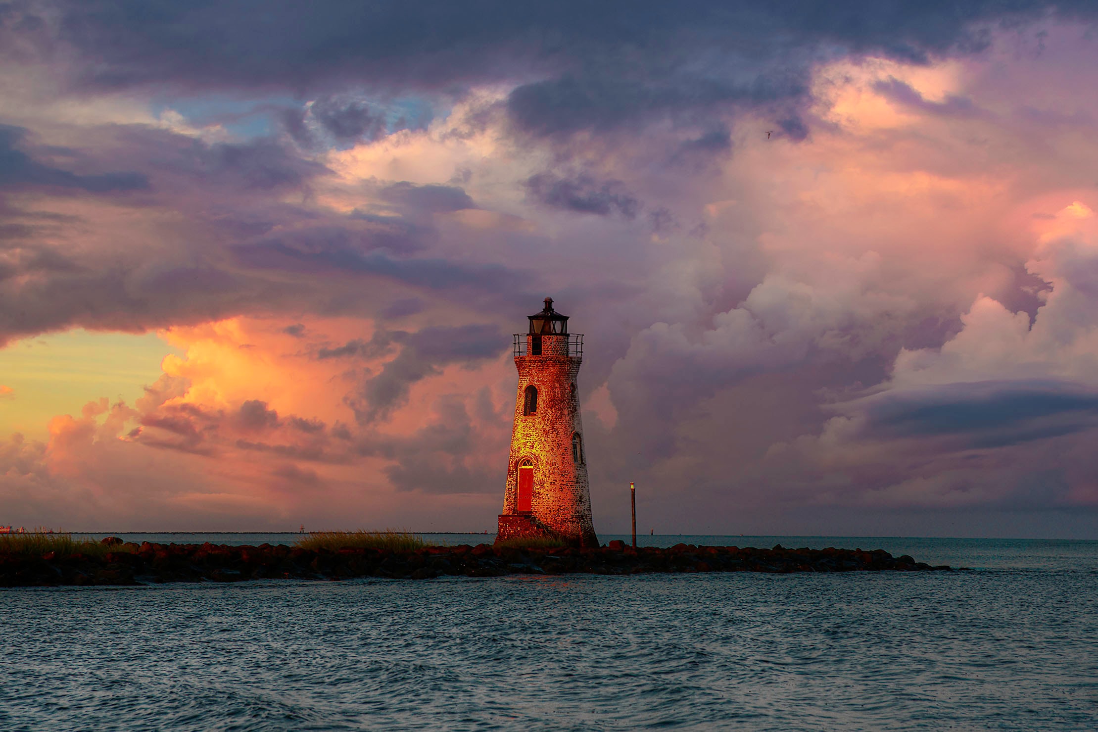 Cockspur Island Lighthouse glowing amber in the last light of sunset beneath a dramatic sky of violet and orange storm clouds
