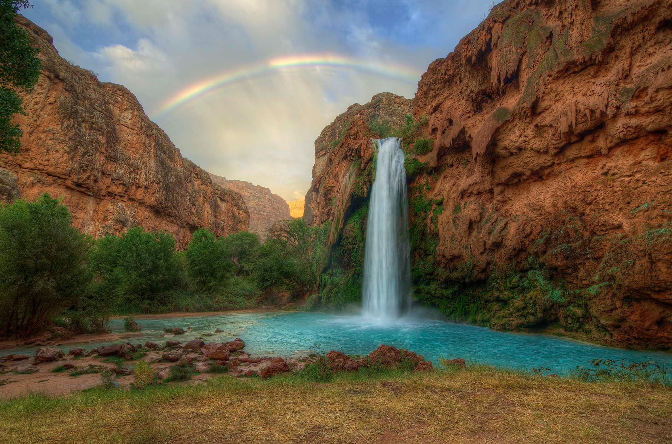 Havasu Falls roaring after a desert rain, turquoise water cascading into a vivid blue-green pool ringed by red canyon walls