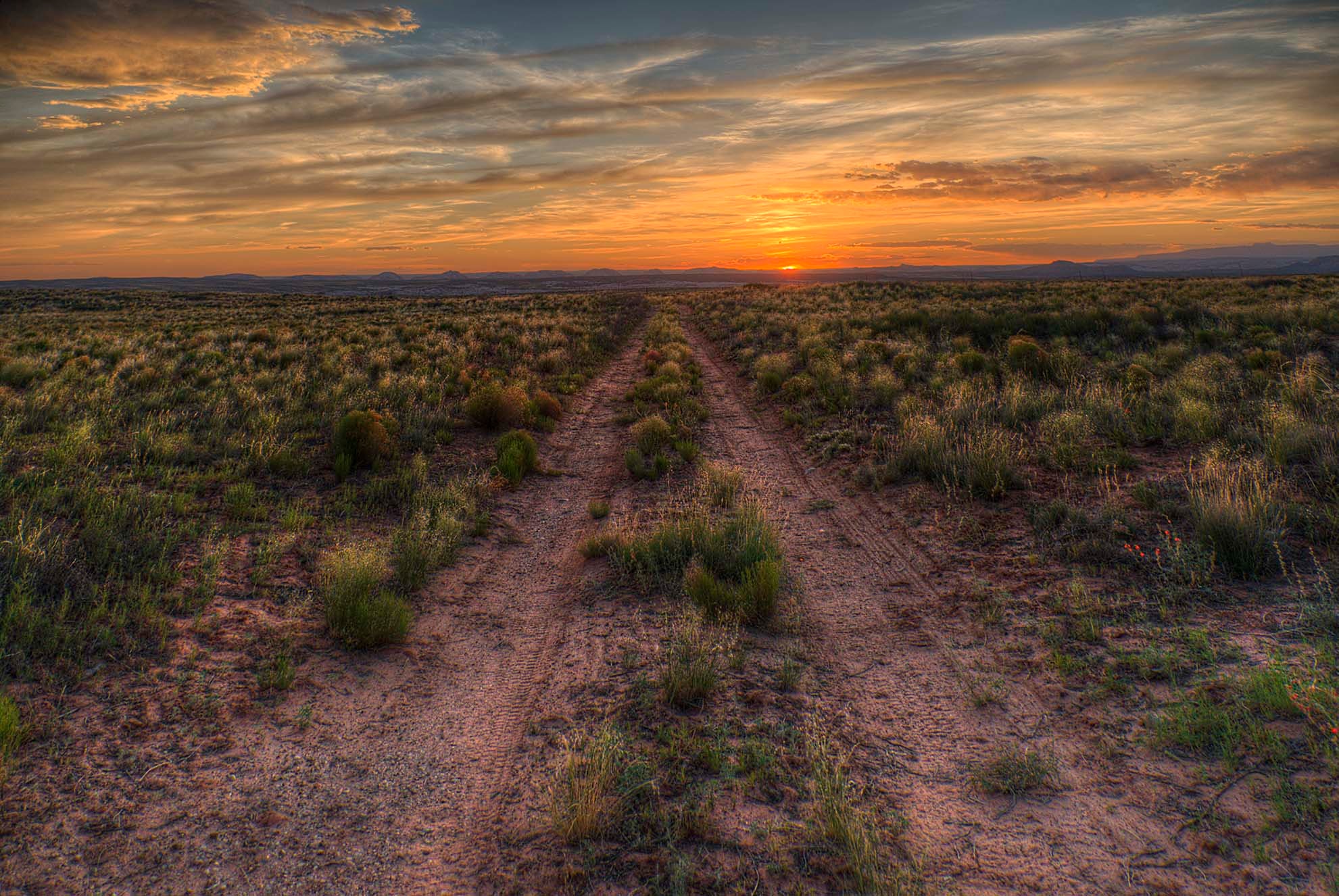 A dirt track disappearing into the open Utah desert as the sun sets in a blaze of orange and gold on the horizon