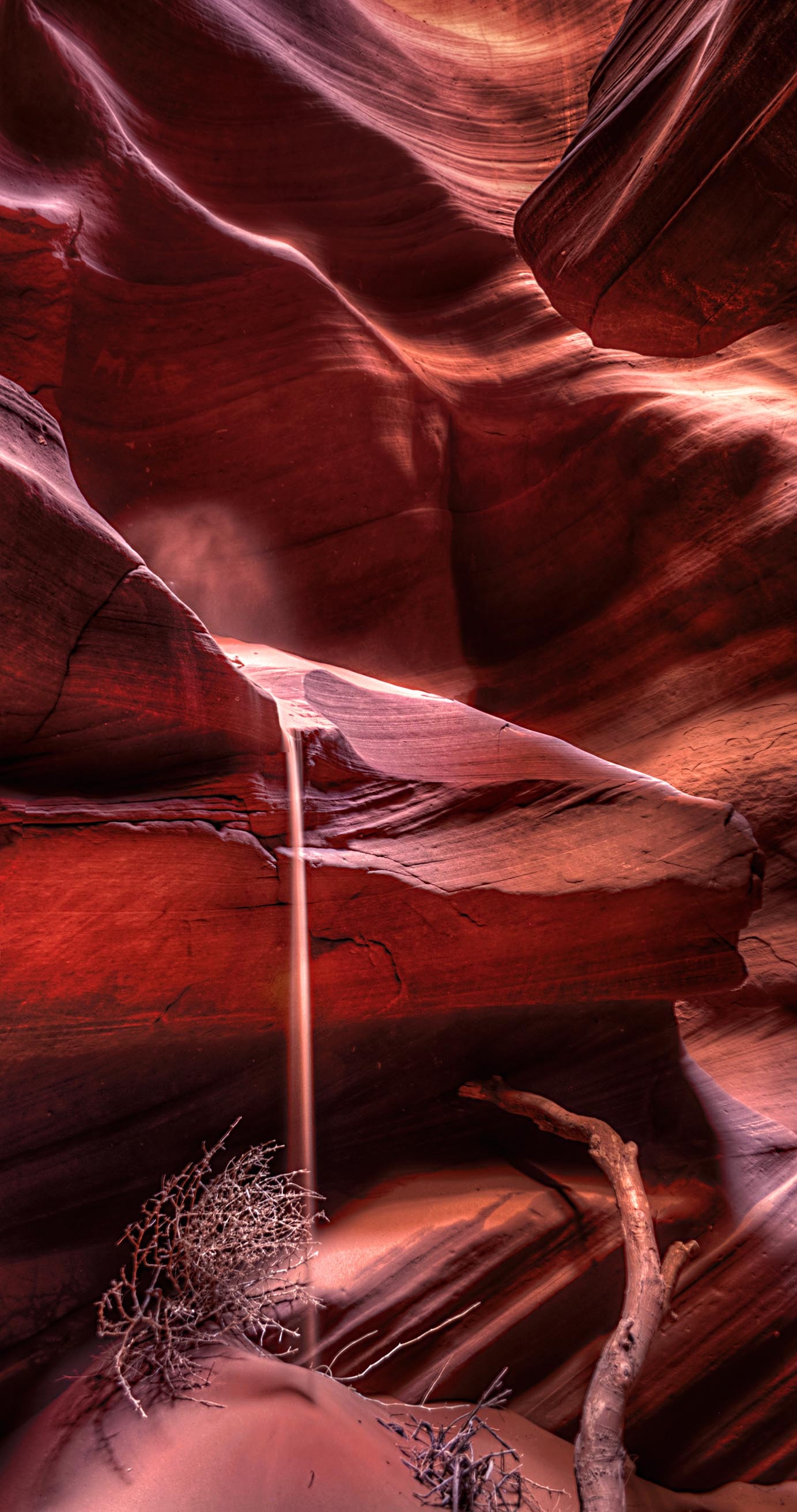 A thin stream of sand falling through a cathedral of sculpted red sandstone deep inside Antelope Canyon, driftwood resting on the canyon floor