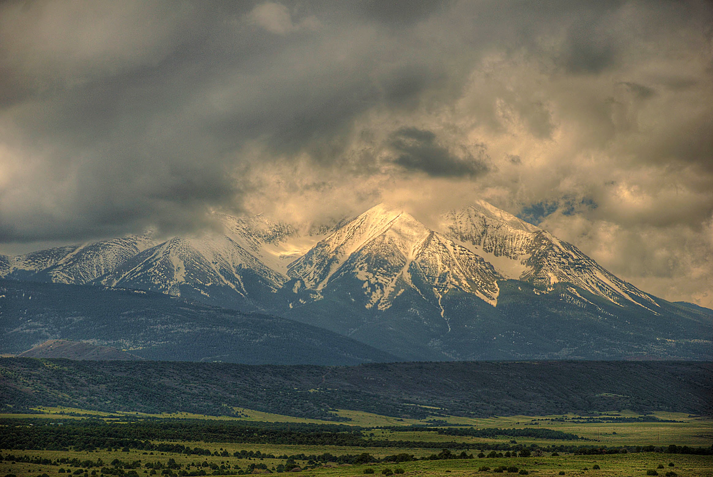 Snow-dusted peaks of the Spanish Peaks emerge from brooding storm clouds above a wide green valley, southern Colorado