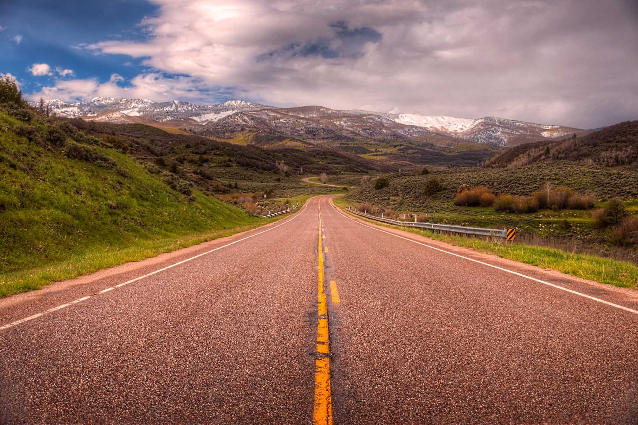 A two-lane highway stretches toward snow-capped Rocky Mountain peaks under a wide blue sky with dramatic clouds, Colorado