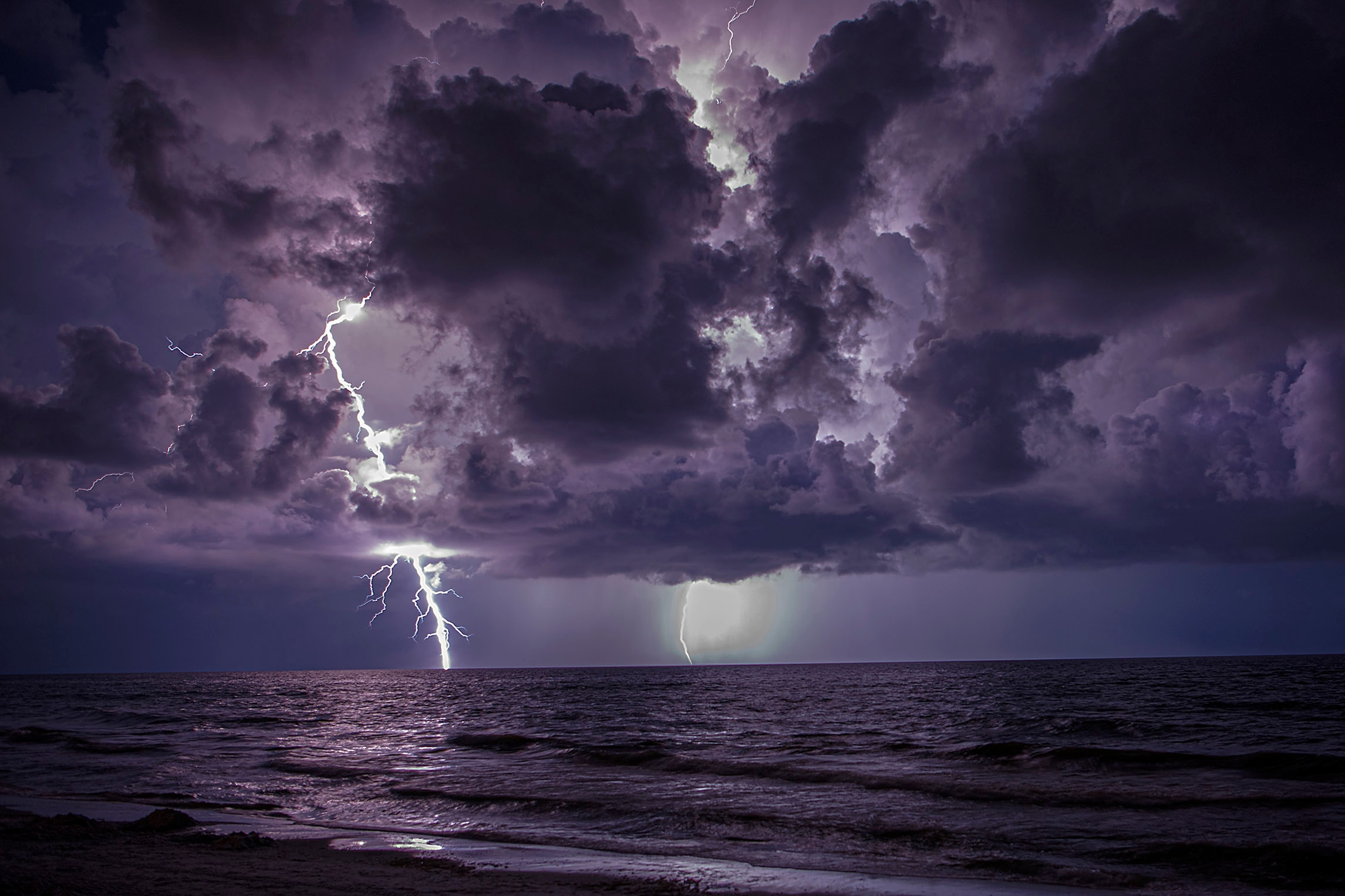 Multiple lightning bolts illuminating a purple storm sky over the ocean, reflected in the calm dark water along a beach