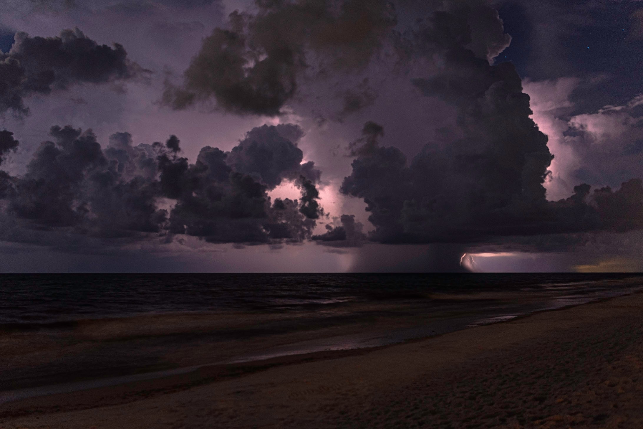 A towering cumulonimbus storm cell lit from within by lightning over a dark calm ocean at night